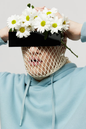 a strange man stands on a light background with a mesh and black glasses covering his eyes, turning his face to the camera holding chamomile flowers on his headの写真素材