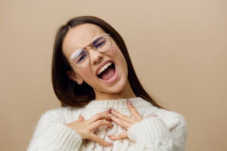 An emotional, loudly screaming woman stands on a beige studio background in a white sweater with her eyes closedの写真素材