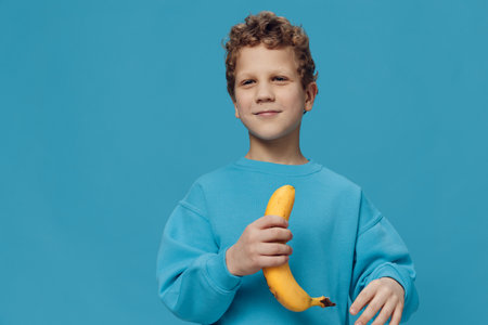a beautiful, happy boy stands in blue clothes on a blue background and holds a banana in his hand smiling joyfully at the camera. Horizontal studio photo with empty space for advertising insertの写真素材