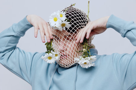 a close horizontal portrait of a strange man standing on a light background in a blue hoodie with a mesh on his face, to which white daisies are attached and poses with his hands relaxed near his faceの写真素材
