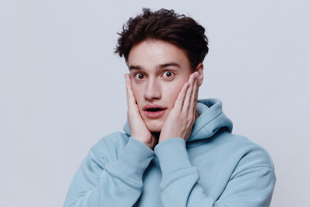 a cute dark-haired young man stands in a light blue hoodie on a light background and smiles...