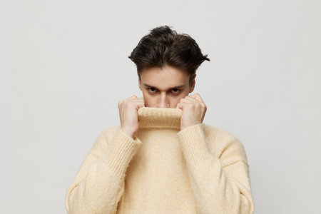 Horizontal studio shot. a cute dark-haired young man stands in a beige turtleneck on a light gray background and smiles pleasantly, pointing his hands in different directions.の写真素材