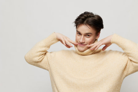 Portrait photo. Horizontal studio shot. close-up photo.A cute young man with dark short curly hair combed back in a beige sweater with a high neck stands on a gray backgroundの写真素材