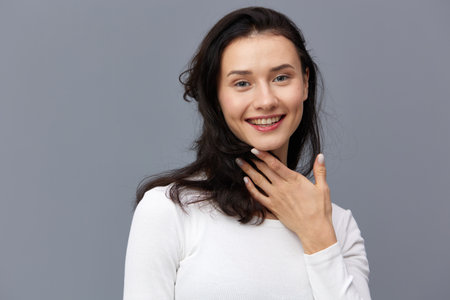 an excited woman stands on a gray background in a white T-shirt, covered her neck with her hair,...