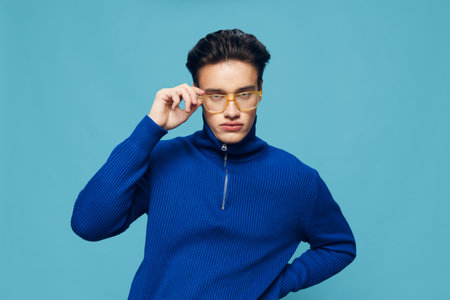 a cute, handsome man stands on a light blue background in a blue zip-up sweater and holds yellow sunglasses with his hand while looking at the camera. Studio photographyの写真素材
