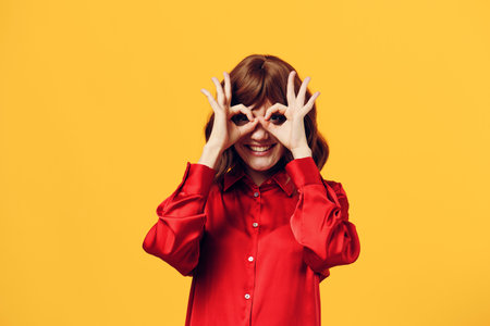 a funny, cheerful woman stands in a stylish red shirt on a yellow background and looking into the camera happily shows the OK sign with her fingers, holding her hands in front of her eyes like a maskの写真素材