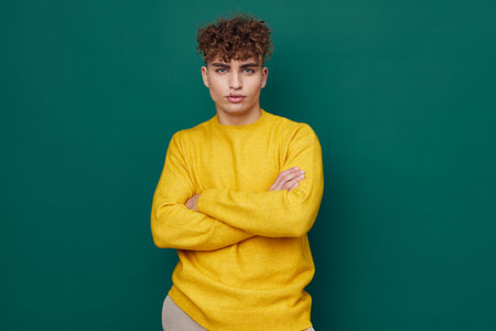 a handsome man stands on a green background in a yellow sweater and smiling pleasantly poses with his arms folded on his chest . Horizontal photo with empty spaceの写真素材