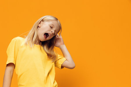 a cute sleepy school-age girl stands on a yellow background and yawns with her mouth wide openの写真素材