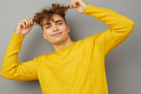 a cute, charming man stands on a gray background, smiling pleasantly at the camera, holding his hand on his hair in a small curl. Horizontal close-up portrait in the studioの写真素材