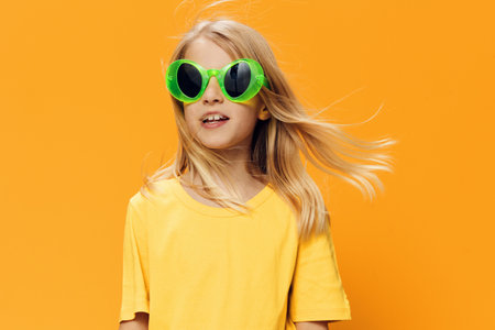 a beautiful girl in funny green sunglasses stands in bright clothes on an orange background spreading her arms to the sidesの写真素材