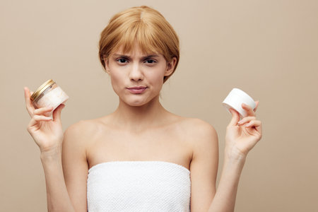 A nice woman in a towel with well-groomed skin, red hair pinned back hair on a beige background with two jars of cream is very happy about a new purchase.Horizontal studio shot.の写真素材