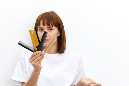 pleasant woman with a beautiful hairstyle holds out a lot of combs in her hand to the camera and looks through them, raising her hand in a relaxed gesture, standing on a white background in a T-shirtの写真素材