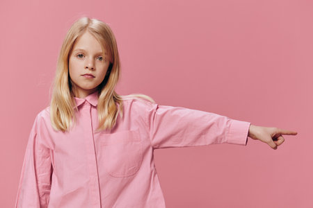 a beautiful, small, funny girl stands in a pink shirt on a pink background and points with her index finger towards the empty space on the background under an advertising mockupの写真素材
