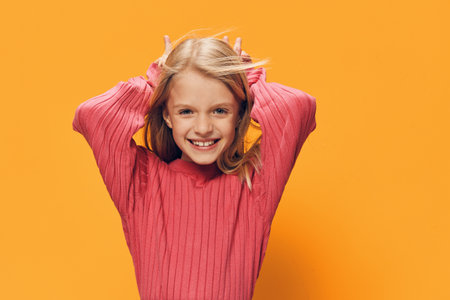 a cute, happy school-age girl stands on a yellow background in a pink sweater with her hands behind her head, smiling broadly at the cameraの写真素材