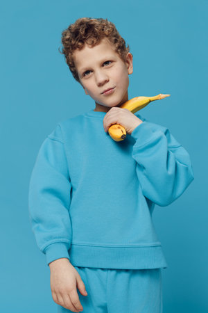 a beautiful, happy boy stands in blue clothes on a blue background and holds a banana in his hand smiling joyfully. Vertical studio photoの写真素材