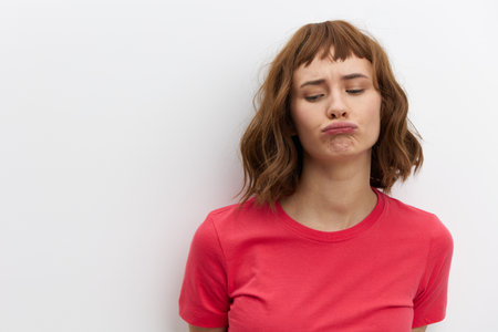 a funny, emotional, slightly upset woman stands in a red cotton T-shirt on a white background and makes a funny face showing her emotionsの写真素材