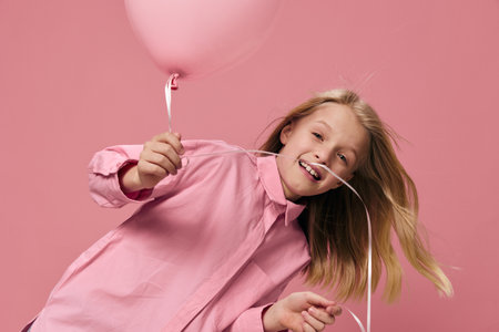 happy, joyful girl of school age, blonde, stands playing with a pink balloon in her hand and smiles joyfullyの写真素材