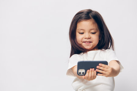 little girl in a white t-shirt stands on a light background with a fashionable smartphone in her...