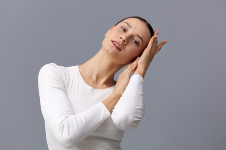 a beautiful, slender, elegant woman stands on a gray background in a white T-shirt with long sleeves and gently touches her neck with her fingers while looking at the camera, smiling pleasantly.の写真素材