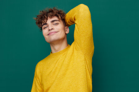 a relaxed man with curly hair, standing on a green background in a yellow sweater and closing his...