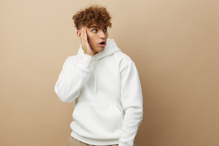 a funny, shocked man stands on a beige background in a white hoodie and holds his hands wide open near his face, expressing emotionsの写真素材
