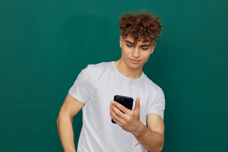 a funny, surprised man with curly hair stands against a green background and holds his smartphone with both hands, carefully looking into it. Horizontal photography in the studioの写真素材