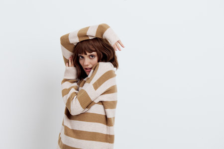 horizontal photo on a light background of a pleasant, beautiful, attractive woman standing in a stylish striped sweater posing with her hands near her face looking into the camera.の写真素材
