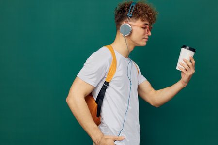 man stands on a green background listening to music with headphones with bright glasses and a backpack on his back holding a cardboard glass for hot drinks in his hand, standing sideways to the cameraの写真素材