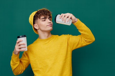 man stands on a green background in a yellow T-shirt and hat holding cardboard glasses for hot drinks in his hand and raises one to his mouth. Horizontal photo for inserting an advertising layoutの写真素材