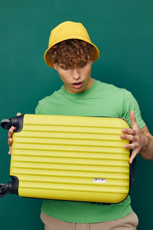 a cute, surprised man stands on a green background, dressed in a yellow panama hat, holding a bright travel suitcase in his hand, his mouth wide openの写真素材