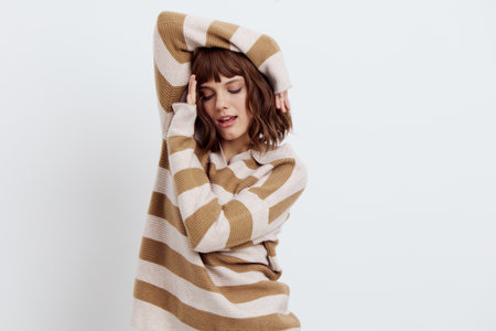 photo on a light background of beautiful, attractive woman standing in a stylish striped sweater, relaxed posing with her hands near her face with her eyes closed. A photo with an empty spaceの写真素材