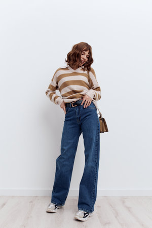 a full-length photo of a beautiful, stylish woman standing on a light background in a striped sweater and blue jeans, posing relaxed with a beige bag slung over her shoulderの写真素材