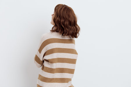 a sweet, pleasant woman with a short haircut stands on a light background in a stylish beige, striped sweater, standing sideways away from the cameraの写真素材
