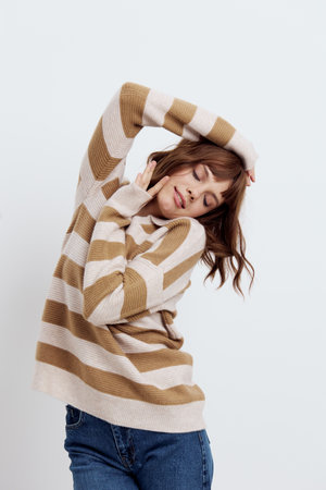 vertical photo of a beautiful, stylish woman in a fashionable striped sweater standing on a light background and posing with her hands near her face with her eyes closedの写真素材