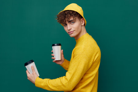 a joyful man stands on a green background in bright clothes and holds two coffee glasses in his...