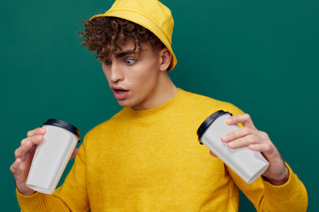 a surprised man stands on an iron background in a yellow T-shirt and hat holding two cardboard glasses in his hands. Horizontal photo for inserting an advertising layoutの写真素材