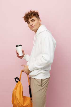 a nice man is standing on a pink background in a white hoodie holding an orange backpack and a cardboard cup looking away. Vertical photographyの写真素材