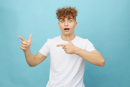 an emotional, surprised man stands on a light blue background with his mouth wide open from emotions, pointing his fingers towards an empty space, attracting attentionの写真素材