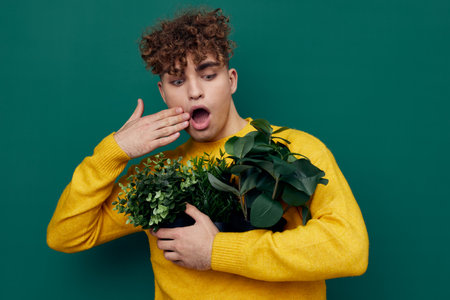 a shocked man stands on a green background in bright clothes holding potted plants in his hands and covers his mouth wide open in surprise with his handの写真素材