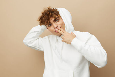 a stylish young man in a white hoodie, standing on a beige background and holding his head with one hand and bringing the other to his lipsの写真素材