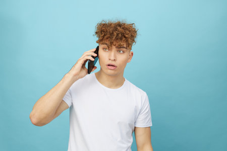 shocked man with curly hair stands on a background and holds his smartphone looking into it with surprise on his face. Horizontal studio photo with an empty space for inserting an advertising layoutの写真素材