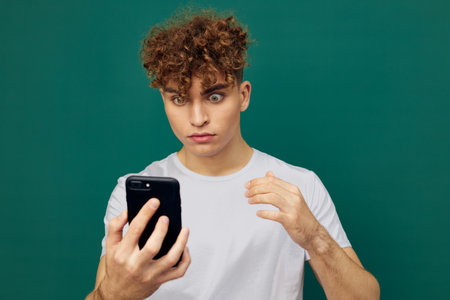 man with curly hair stands against green background and holds his smartphone looking into it with surprise on his face. Horizontal studio photo with an empty space for inserting an advertising layoutの写真素材