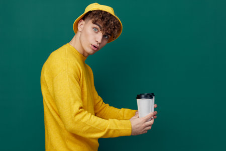 a joyful man stands on a green background in bright clothes and holds two coffee glasses in his hands, smiling affably at the cameraの写真素材