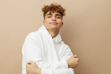 an attractive man stands on a beige background in a white hoodie and looking at the camera smiles pleasantly. Horizontal photo with an empty space for inserting an advertising layoutの写真素材