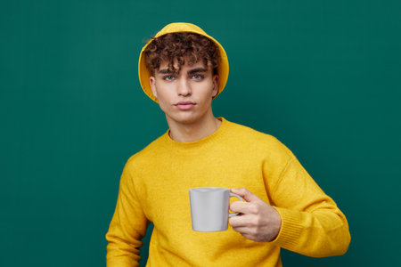 a handsome man stands on a green background in a yellow T-shirt and hat holding a metal glass for hot drinks in his hand and looks at the camera. Horizontal photo for inserting an advertising layoutの写真素材