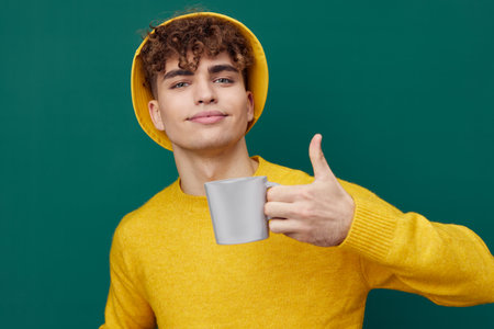 a handsome man stands on a green background in a yellow T-shirt and hat holding a metal glass for hot drinks in his hand and looks at the camera. Horizontal photo for inserting an advertising layoutの写真素材