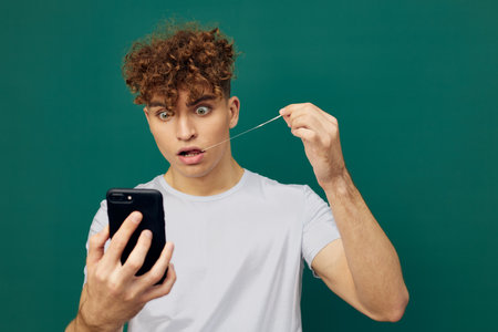 a funny, shocked man with curly hair stands against a green background and holds his smartphone looking into it with surprise on his face, chewing gum and pulling it out of his mouth to the side.の写真素材
