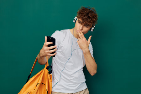 a joyful man, a teenager, stands on a green background listening to music with headphones, holding his smartphone in his hand and looking at it, touching his mouth with his fingersの写真素材