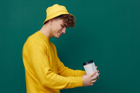 a joyful man stands on a green background in bright clothes and holds two coffee glasses in his hands, smiling affably and looking downの写真素材