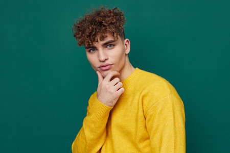 a cute, thoughtful man stands on a green background in a yellow sweater holding his hand to the will of the face. Horizontal photo with empty spaceの写真素材